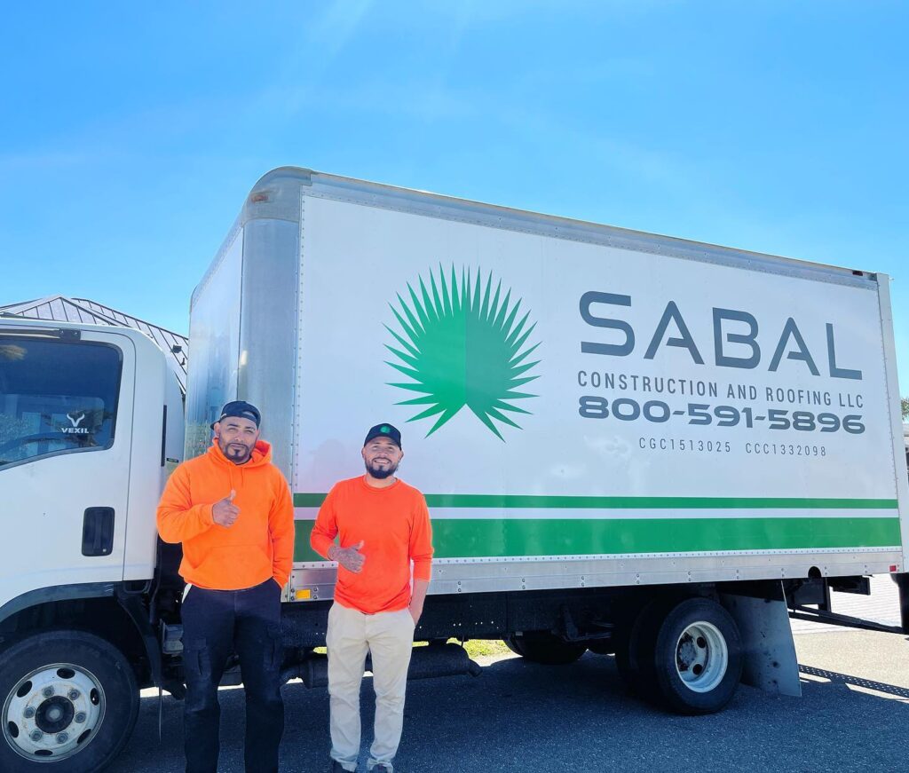 Two men in orange shirts are standing in front of a truck, giving a thumbs up